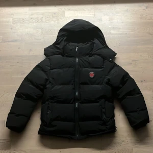 Svart pufferjacka från Trapstar - Säljer en svart pufferjacka från Trapstar med stor huva och dragkedja framtill. Jackan har en broderad logga på bröstet och TRAPSTAR i rött på ryggen. Fylld för extra värme och har två fickor framtill. Perfekt för kalla dagar.