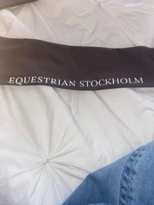 Svart långärmad topp Equestrian Stockholm - Snygg svart långärmad topp från Equestrian Stockholm med vit logga längs ena ärmen. Figurnära passform och tillverkad i ett stretchigt syntetmaterial som passar perfekt till ridning eller träning.