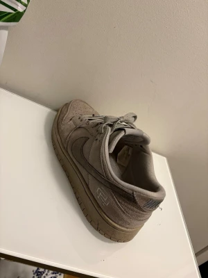 Nike Dunk Low beige mocka sneakers - Säljer ett par Nike Dunk Low sneakers i beige mocka med matchande snören och gummisula. Skorna har klassisk swoosh på sidan och diskreta detaljer. Perfekta för dig som gillar stilrena och neutrala färger.