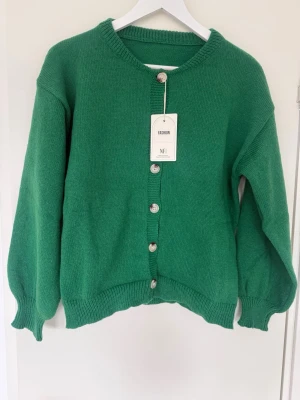 Grön stickad kofta med pärlemorknappar - Snygg grön stickad kofta från NFI med rund halsringning och pärlemorskimrande knappar framtill. Koftan har ribbade muddar vid ärmslut och nederkant samt en rak, avslappnad passform. Perfekt att slänga över en t-shirt eller topp.