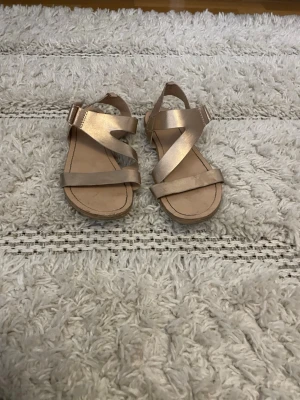 Guldiga sandaler med remmar - Snygga sandaler i metallic guld med breda korsade remmar och justerbart spänne vid ankeln. Tillverkade i konstläder med platt sula och öppen tå. Perfekta för sommaren och ger en trendig touch till din outfit. Använda men ändå väldigt bekväma med ett bra skick 