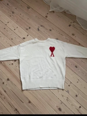 Vit stickad tröja från Ami Paris - Vit stickad tröja från Ami Paris med röd hjärtlogga och bokstaven 'A' på bröstet. Tröjan har rund halsringning, ribbade muddar och är tillverkad i mjukt material. Perfekt för dig som gillar stilrena plagg med en twist.