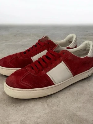 Valentino Flycrews - Snygga röda sneakers från Valentino med vit sidopanel och vit sula. Ovandelen är i rött mocka och skorna har matchande röda skosnören. Klassisk låg modell med rund tå och diskret logga på plösen. Perfekta för dig som vill sticka ut med stil.