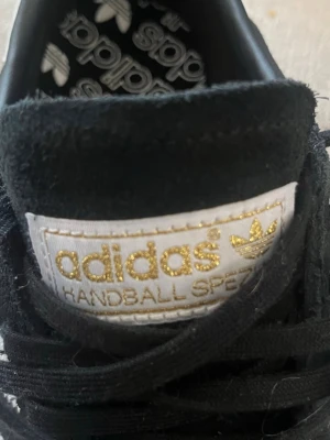 Adidas Handball Spezial svart/vit - Säljer ett par Adidas Handball Spezial sneakers i svart mocka med vita detaljer och gummisula. Klassisk Adidas-logga i guld på plösen och retroinspirerad design. Skorna har svarta snören och en låg silhuett som passar till många stilar.