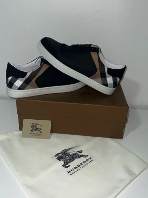 Burberry svartvita sneakers - Snygga sneakers från Burberry med klassiskt rutmönster i beige, svart och vitt på sidorna. Ovandelen är svart med vita sulor och svarta skosnören. Materialet är en mix av textil och läderdetaljer. Perfekta för dig som vill ha en stilren och trendig look.