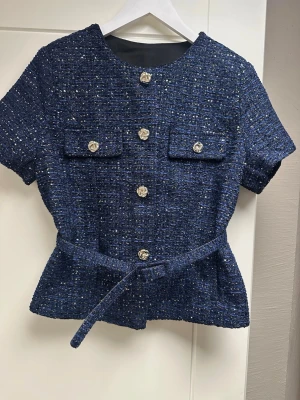 Mörkblå tweedblus med bälte S - Stilren mörkblå tweedblus med korta ärmar, dekorativa guldknappar och två bröstfickor. Blusen har ett matchande bälte i midjan som ger en snygg siluett. Materialet är strukturerat och har inslag av vitt och guld för en exklusiv känsla.