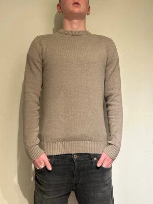 Beige stickad tröja i klassisk modell - Snygg beige stickad tröja med rund halsringning och ribbade muddar vid ärmslut och nederkant. Enkel och stilren design som passar till det mesta. Perfekt för lager-på-lager och enkel att matcha med jeans eller chinos.
