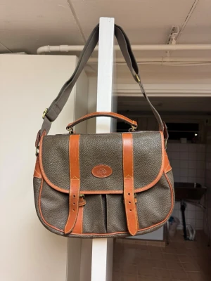 Mulberry axelväska i läder – klassisk modell - Säljer en äkta Mulberry axelväska i tidlös modell. Väskan är i mörkbrunt/grönt kornigt läder med bruna läderdetaljer och justerbar axelrem i textil/läder. Klassisk Mulberry-design med spännen framtill och logodetaljer.  Detaljer: 	•	Märke: Mulberry 	•	Tillverkad i England 	•	Material: Äkta läder 	•	Färg: Mörkgrön/brun med bruna detaljer 	•	Justerbar axelrem 	•	Flera innerfack + ytterfack 	•	Stängs med spänne  Skick: Använd men i gott vintage-skick.   Perfekt vardagsväska eller för dig som gillar