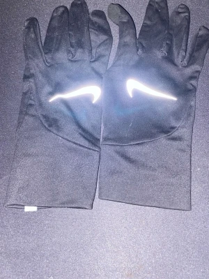 Svarta Nike träningshandskar - Säljer ett par svarta träningshandskar från Nike med den klassiska vita swoosh-loggan på ovansidan. Handskarna är tunna och smidiga, perfekta för löpning eller utomhusträning. Tillverkade i ett stretchigt syntetmaterial som sitter skönt på handen.
