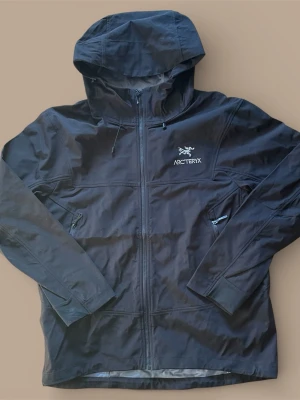 Svart vindjacka från Arc'teryx - Svart vindjacka från Arc'teryx med huva och dragkedja framtill. Jackan har justerbar dragsko vid huvan och logga på bröstet. Perfekt för blåsiga dagar och enkel att matcha med olika outfits. Det står stolek L men passar bättre som storlek M.