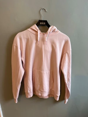 Ljusrosa hoodie med huva - En stilren ljusrosa hoodie med huva och dragsnören. Hoodien har en stor magficka framtill och ribbade muddar vid ärmslut och nederkant.  Tillverkad i mjukt bomullstyg. Använd ca två gånger. Kan absolut gå med på att ändra pris