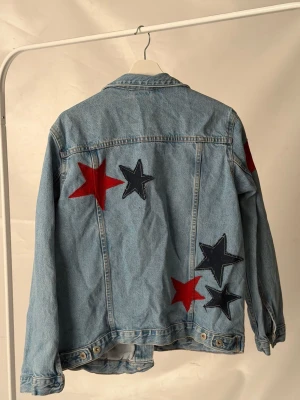 Jeansjacka med stjärnor - Jeansjacka med stora röda och svarta stjärnapplikationer på ryggen och ena ärmen. Perfekt för dig som vill sticka ut med en unik detalj i din outfit.🩵