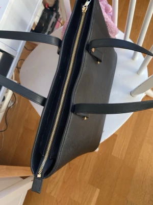 Svart handväska med gulddetaljer - Stilren svart handväska i fyrkantig modell med två långa handtag och guldfärgade metalldetaljer. Väskan har dragkedja upptill och små metallfötter undertill. Materialet är slätt och tåligt, perfekt för dig som gillar klassisk och enkel design.