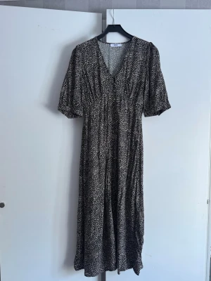 Svartvit mönstrad midiklänning - Svartvit midiklänning med små abstrakta prickar, v-ringning och korta puffärmar. Klänningen har knappar framtill och en slits nertill för extra rörelsefrihet. Tillverkad i ett lätt och luftigt tyg. 