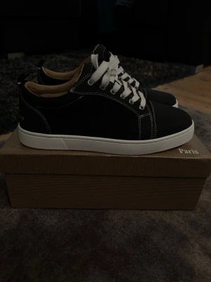 Svarta Louboutin sneakers med röd sula - Snygga svarta sneakers från Christian Louboutin med klassisk röd sula och vita kontrastsömmar. Skorna har vita snören och en låg silhuett. Ovandelen är i mocka och insidan är beige. Perfekta för dig som vill sticka ut med exklusiv stil.