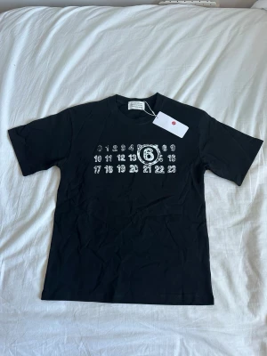 Svart t-shirt från Maison Margiela - Cool svart t-shirt från Maison Margiela med deras ikoniska siffror tryckta i vitt på bröstet. Klassisk passform med rund hals och korta ärmar. Perfekt för dig som gillar stilrena och unika plagg med designer-touch.