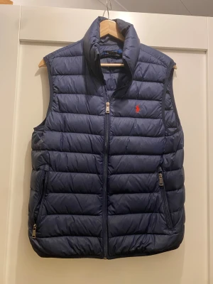 Mörkblå dunväst från Polo Ralph Lauren - Snygg mörkblå dunväst från Polo Ralph Lauren med quiltad design och hög krage. Västen har två dragkedjeförsedda fickor och en röd broderad logga på bröstet. Perfekt att slänga över en hoodie eller tröja för extra värme.