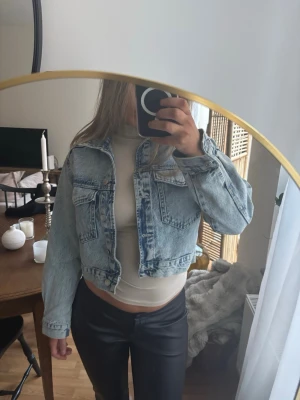 Cropped ljusblå jeansjacka - Trendig cropped jeansjacka i ljusblå denim med två bröstfickor och klassisk krage. Jackan har en avslappnad passform och silverfärgade knappar framtill. Perfekt att slänga över en topp för en cool och avslappnad look.
