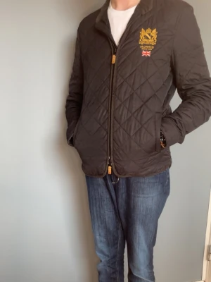 Morris Jacka - Quiltad svart jacka från Morris med guldfärgat broderi och brittisk flagga på bröstet. Jackan har dragkedja framtill, två fickor och klassisk krage. Perfekt för dig som gillar stilren och tidlös design.