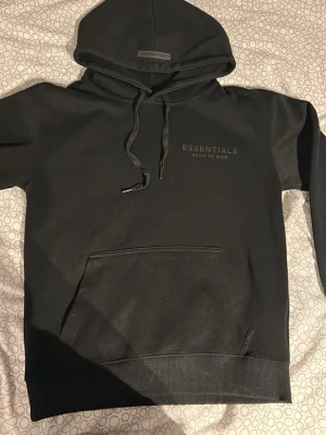 Svart Essentials Fear of God hoodie - Svart hoodie från Fear of God Essentials med tryckt logga på bröstet och ryggen. Klassisk känguruficka framtill, ribbade muddar och justerbar dragsko i huvan. Perfekt för en avslappnad och trendig streetwear-look.