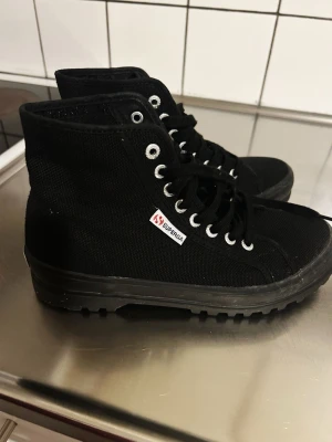 Superga Alpina 996 Full Black sneakers - Snygga svarta Superga Alpina 996 sneakers med högt skaft och grov sula. Skorna är tillverkade i canvas med svarta skosnören och silverfärgade öljetter. Klassisk Superga-logga på sidan. Perfekta för dig som gillar streetstyle och vill ha något som sticker ut.