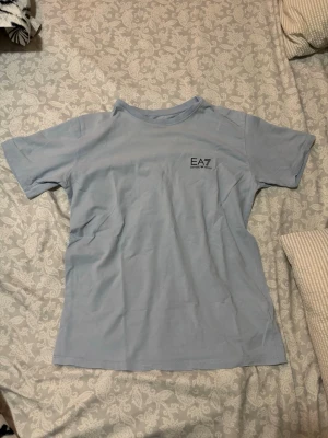 Ljusblå EA7 t-shirt i bomull - Snygg ljusblå t-shirt från EA7 Emporio Armani med diskret logga på bröstet. Klassisk rund hals och korta ärmar. Tillverkad i mjuk bomull som känns skön mot huden. Perfekt för en clean och stilren look.