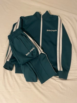 Grönt tracksuit set från Palm Angels - Snyggt grönt tracksuit set från Palm Angels med vita ränder längs ärmar och ben. Setet består av en zip-jacka med hög krage och matchande byxor med resår i midjan. Båda delarna har Palm Angels-loggan tryckt i vitt. Materialet är troligtvis polyester för en sportig känsla.