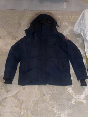 Mörkblå dunjacka från Canada Goose - Mörkblå dunjacka från Canada Goose med huva och pälskant. Klassisk modell med logotyp på ärmen, stora fickor och dragkedja med knappar. Perfekt för kalla vinterdagar och riktigt varm.