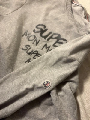 Grå sweatshirt från Moncler - Snygg grå sweatshirt från Moncler med tryck i svart och vitt på bröstet där det står 'SUPEEER MON MONCLER!'. Tröjan har rund halsringning, ribbade muddar och en liten Moncler-logga på ärmen. Perfekt för en avslappnad och trendig look.