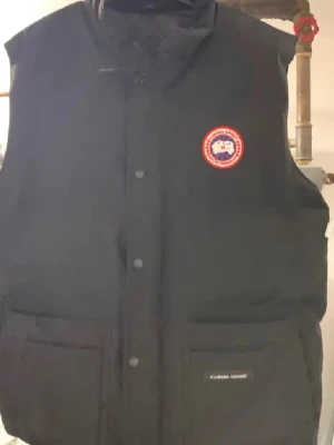 Svart dunväst från Canada Goose - Säljer en svart dunväst från Canada Goose med klassisk logotyp på bröstet och två stora fickor framtill. Västen har hög krage och stängs med både dragkedja och tryckknappar. Perfekt för lager-på-lager och riktigt varm.