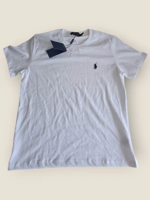 Vit t-shirt från Polo Ralph Lauren - Klassisk vit t-shirt från Polo Ralph Lauren med den ikoniska broderade loggan på bröstet. T-shirten har rund halsringning och korta ärmar. Tillverkad i mjuk bomull som känns skön mot huden. Perfekt basplagg till garderoben.