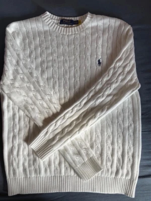 Vit kabelstickad tröja Polo Ralph Lauren - Kabelstickad vit tröja från Polo Ralph Lauren med rund halsringning och klassisk marinblå logga broderad på bröstet. Tröjan har ribbade muddar vid ärmslut och nederkant. Perfekt för dig som gillar stilrena och tidlösa plagg.