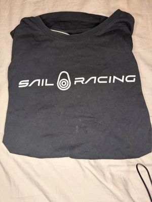 Svart Sail Racing t-shirt - Säljer en svart t-shirt från Sail Racing med vit logga tryckt över bröstet. Klassisk rund hals och korta ärmar. Tillverkad i mjuk bomull som känns skön mot huden. Perfekt för dig som gillar stilrena och sportiga plagg.