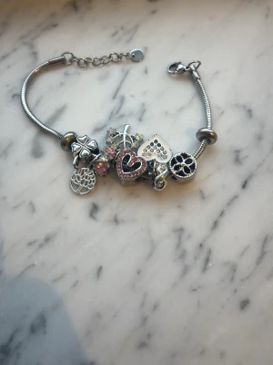 Silverfärgat charmarmband  - Säljer ett silverfärgat charmarmband med flera berlocker, bland annat hjärtan, fyrklöver och kors. Några av berlockerna har gnistrande stenar och rosa detaljer. Armbandet har en kedja och är perfekt för dig som gillar personliga smycken med många detaljer. 