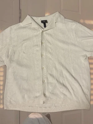 Vit ribbad skjorta från H&M - Vit skjorta från H&M med korta ärmar och klassisk krage. Skjortan har ribbad struktur och knäppning framtill. Perfekt för en clean och stilren look.