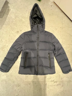 Dunjacka J.lindeberg - Snygg mörkgrå pufferjacka från J.Lindeberg med huva och dragkedja framtill. Jackan har två stora fickor med dragkedja och quiltad design som håller värmen. Perfekt för kalla dagar och har en stilren look som passar till det mesta. Har gjorts en liten lagning på ärmen (se bild) men annars är den i riktigt bra skick. Vid frågor eller funderingar är det bara att höra av sig!           Nypris~4500.                                                                             Mitt pris: 2000