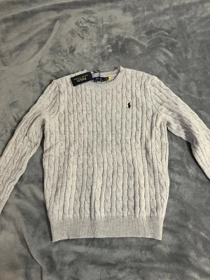 Ljusgrå kabelstickad tröja Polo Ralph Lauren - Kabelstickad ljusgrå tröja från Polo Ralph Lauren med rund halsringning och klassisk svart broderad logga på bröstet. Tröjan har ribbade muddar och nederkant, perfekt för en stilren och tidlös look.