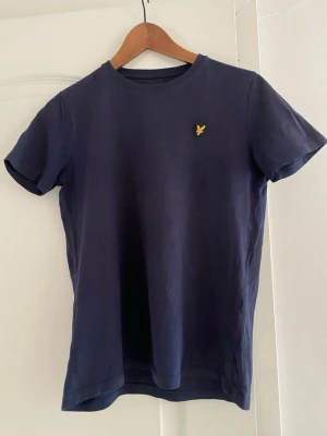 Mörkblå t-shirt från Lyle & Scott - Klassisk mörkblå t-shirt från Lyle & Scott med rund hals och korta ärmar. T-shirten har den ikoniska gula loggan broderad på bröstet. Tillverkad i mjuk bomull för en bekväm känsla och enkel stil.