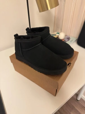 Svarta uggs - Ett par svarta uggs i mocka med mjukt foder på insidan. Skorna har en låg, rundad siluett och grov sula för extra komfort. Perfekta för kalla dagar och ger en avslappnad look. Klassisk design .