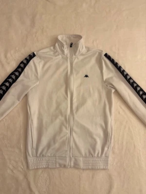 Vit track jacket från Kappa - Vit track jacket från Kappa med svarta ränder längs ärmarna och Kappas logga i vitt. Jackan har dragkedja framtill, hög krage och ribbade muddar vid ärmslut och midja. Perfekt för en sportig och trendig look. Kan gå ner i pris vid snabb affär