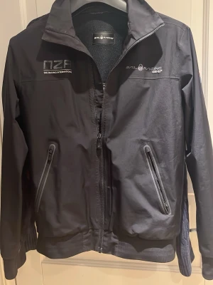  Sail Racing Gore-Tex jacka - Snygg svart vindjacka från Sail Racing med Gore-Tex-material. Jackan har två snedställda dragkedjefickor framtill, hög krage och logga på bröstet. Perfekt för blåsiga dagar och sportig stil. Vattenavvisande och lätt att bära.