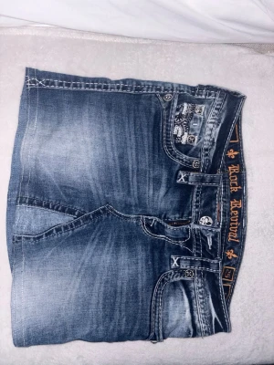 Jeanskjol från Rock Revival - Snygg jeanskjol från Rock Revival i blå denim med slitningar och dekorativa bakfickor med broderade detaljer. Klassisk femficksmodell med knapp- och dragkedjestängning. Perfekt för dig som gillar en cool och avslappnad stil.