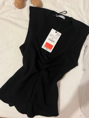 Svart ärmlös topp från Gina Tricot - Snygg svart ärmlös topp från Gina Tricot med v-ringning och knytband i nacken. Toppen har en figurnära passform och är tillverkad i ett mjukt material som känns bekvämt mot huden. Perfekt att styla med jeans eller kjol.