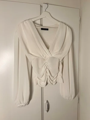 Vit omlottblus från Pastelier - Trendig vit omlottblus från Pastelier med djup v-ringning och rynkade detaljer framtill. Blusen har långa puffärmar med resår vid ärmslut och smockad rygg för en bekväm passform. Tillverkad i ett lätt, flowy tyg som ger en elegant look.