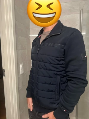 Mörkblå bomberjacka från Tommy Hilfiger - Snygg mörkblå cardigan från Tommy Hilfiger med quiltad front och dragkedja. Jackan har ribbade muddar, ståkrage och diskreta logodetaljer på ärm och handled. Perfekt för dig som vill ha en stilren och bekväm jacka.