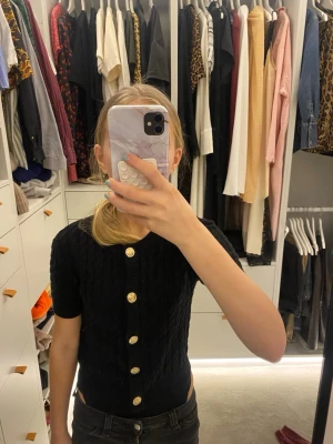 Svart kortärmad kofta med guldknappar - Snygg svart kofta med korta ärmar och dekorativa guldknappar framtill. Koftan har ett kabelstickat mönster och rund halsringning. Perfekt att bära öppen eller stängd för en trendig look.