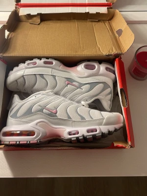 Nike Air Max Plus vita och rosa sneakers - Nike Air Max Plus sneakers i vitt mesh med rosa detaljer och synliga Air-enheter. Swoosh-logga i rosa på sidan och vågiga linjer i designen. Sulan är vit med rosa och svarta inslag. Perfekta för dig som gillar sportig och trendig stil.