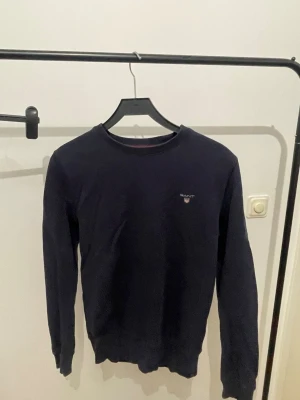 Mörkblå tröja från Gant - Klassisk mörkblå tröja från Gant med rund halsringning och diskret logga broderad på bröstet. Tröjan har långa ärmar och ribbade muddar vid ärmslut och nederkant. Perfekt basplagg för en stilren look.