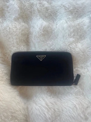 Svart plånbok från Prada - Snygg svart plånbok från Prada i klassisk rektangulär modell med dragkedja runtom. Plånboken har Pradas ikoniska metallogo framtill och är tillverkad i ett tåligt material som ger en lyxig känsla. Perfekt för dig som gillar stilrena accessoarer. Mycket fint skick! Nypris ca 5000kr
