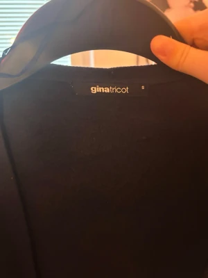 Svart kofta från Gina Tricot - Stilren svart kofta från Gina Tricot med långa ärmar och knytband i midjan. Perfekt att slänga över en topp för en enkel och snygg look. Koftan har en klassisk passform och är tillverkad i ett mjukt material.